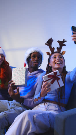 Vertical-Video-Studio-Shot-Of-Gen-Z-Friends-At-Christmas-Sitting-On-Sofa-Wearing-Santa-Hat-And-Reindeer-Antlers-Taking-Selfie-On-Mobile-Phone-2